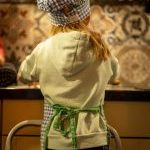 A young girl helping in the kitchen, standing on kitchen stairs, wearing a chef’s hat and apron, seen from the back.