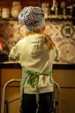 A young girl helping in the kitchen, standing on kitchen stairs, wearing a chef’s hat and apron, seen from the back.